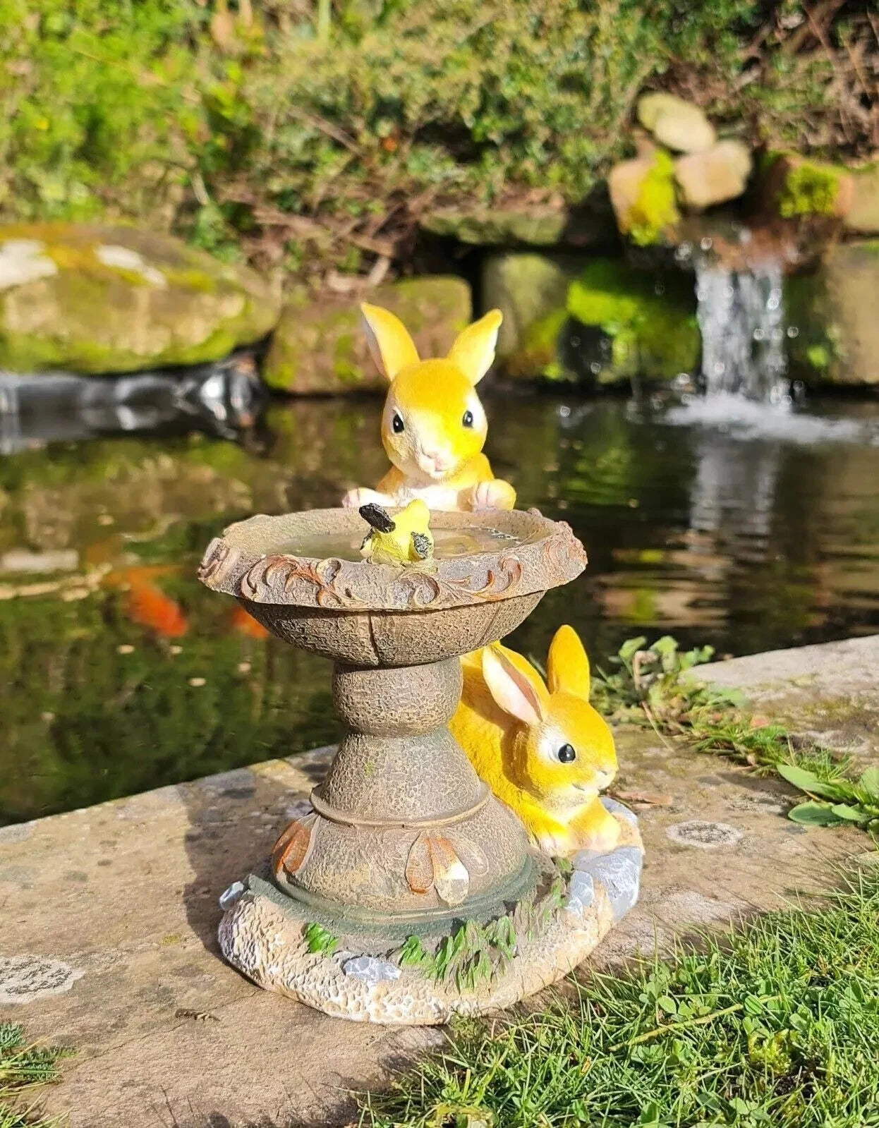 Two yellow rabbit figurines on a stone pedestal by a pond with waterfalls.