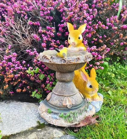 Two yellow rabbit statues on a stone pedestal with pink flowers in the background